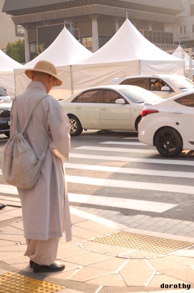 A person standing at a Zebra crossing. The other side of the road has tents put up. There are cars on the road.